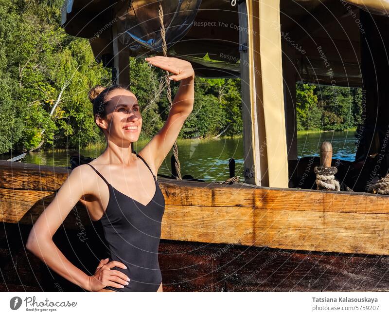 Schlanke junge Frau im Badeanzug, die neben einem alten Holzboot steht und ihr Gesicht mit der Handfläche vor der Sonne schützt schlanke Stehen hölzern Boot