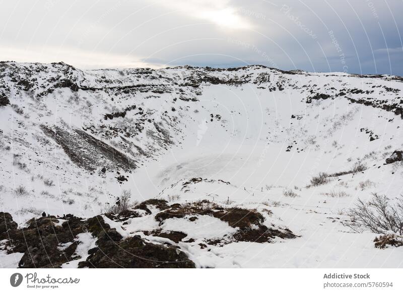 Ein weitläufiger Blick auf einen schneegefüllten Krater in Island, mit zerklüftetem Gelände rund um die eisige Senke unter einem bewölkten Himmel verschneite