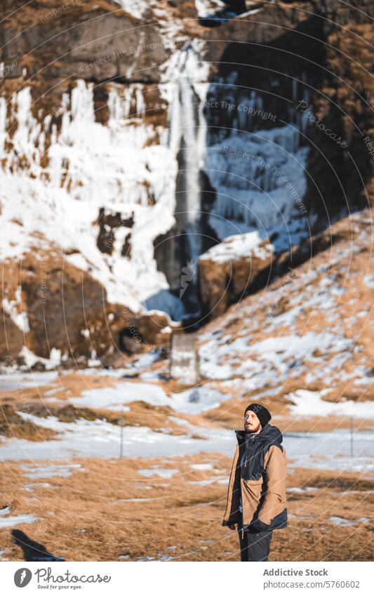 Ein einsamer Wanderer steht nachdenklich vor einem isländischen Wasserfall inmitten der kontrastreichen Winterlandschaft und genießt dessen Erhabenheit Mann