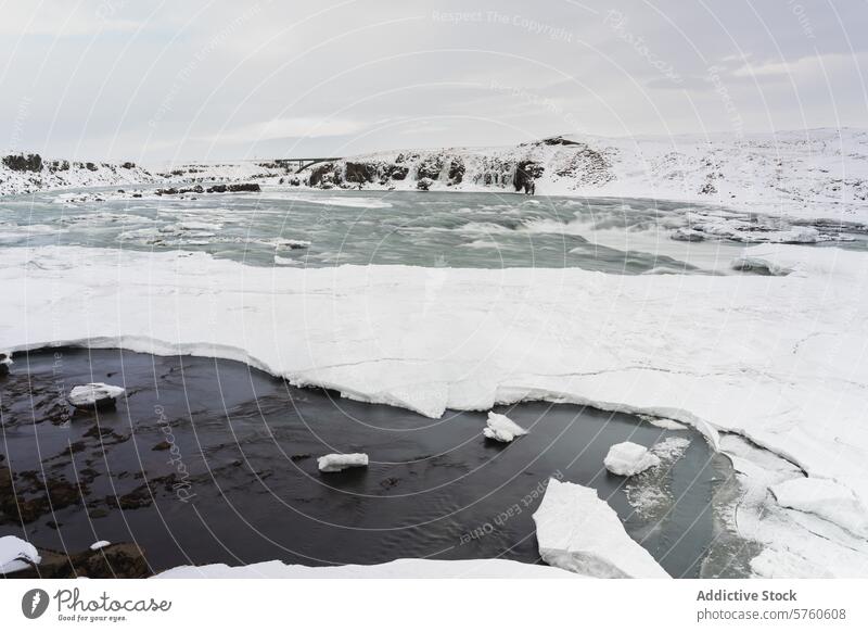 Die schlichte Schönheit eines teilweise zugefrorenen Flusses, der sich unter einem bedeckten Winterhimmel durch die schneebedeckte isländische Landschaft schlängelt