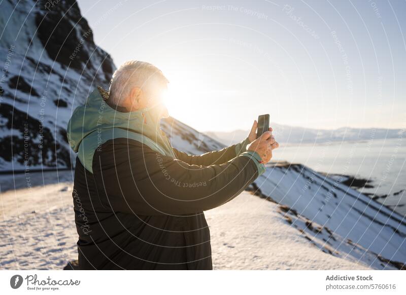 Ein Mann hält den Moment mit seinem Handy fest, als die Wintersonne die verschneite Küste Islands in ein strahlendes Licht taucht erfassend Sonne isländisch