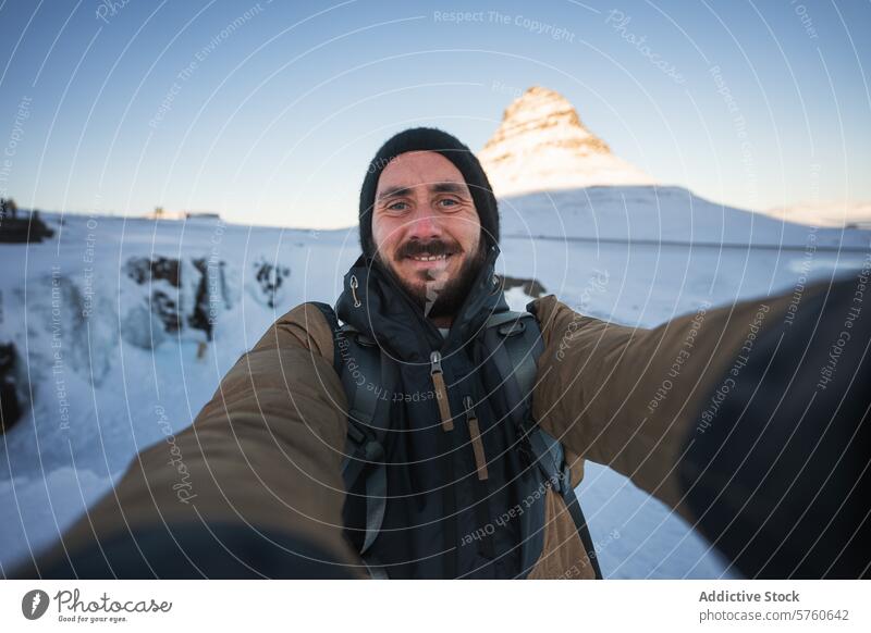 Ein bärtiger Reisender macht ein Selfie mit dem sonnenbeschienenen Berg Kirkjufell, einem berühmten isländischen Wahrzeichen, im verschneiten Hintergrund