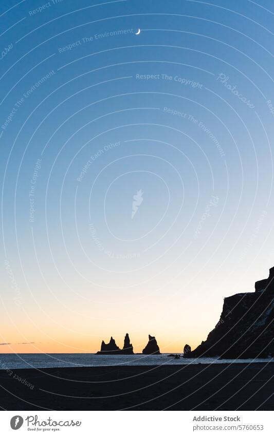 Im Licht der Mondsichel heben sich die Silhouetten der Reynisdrangar-Säulen am schwarzen Sandstrand von Island vom Dämmerungshimmel ab. Gelassenheit Klippen