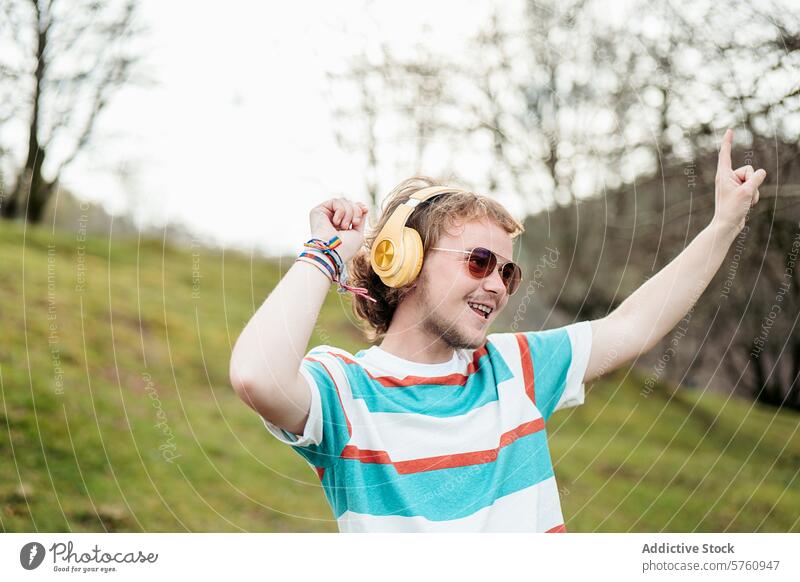 Ein fröhlicher Mann mit breitem Lächeln und gelben Kopfhörern hebt freudig die Hand, während er in einer heiteren Umgebung im Freien tanzt Musik Tanzen Feier