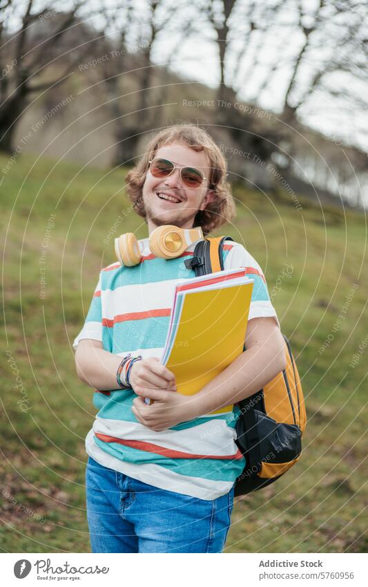 Eine fröhliche Studentin mit lockigem Haar und Sonnenbrille, die ein Notizbuch in der Hand hält und Kopfhörer trägt, steht mit einem Rucksack im Freien Schüler