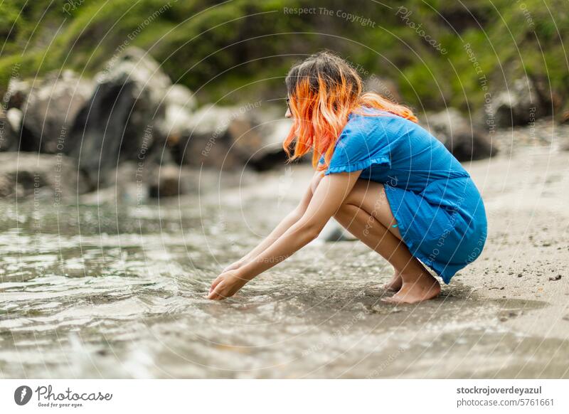 Ein junges Mädchen mit orangefarbenen Haaren, das sich hinhockt und mit den Händen das Wasser berührt, während es auf das Meer blickt Strand Person Sommer MEER