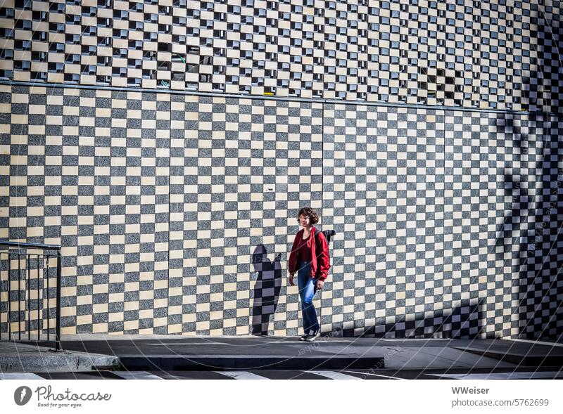 Vor einer schachbrettartig gemusterten Wand läuft ein junges Mädchen die Straße entlang junge Frau Schachbrettmuster Karos Ziegel Rechteck Fassade Mauer
