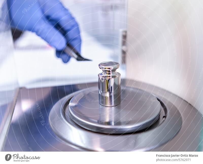 Die Hand des Bedieners hält ein Kalibrierungsgewicht aus Stahl, das auf die Analysenwaage gelegt wird. Konzept der Qualitätskontrolle in einem Labor.
