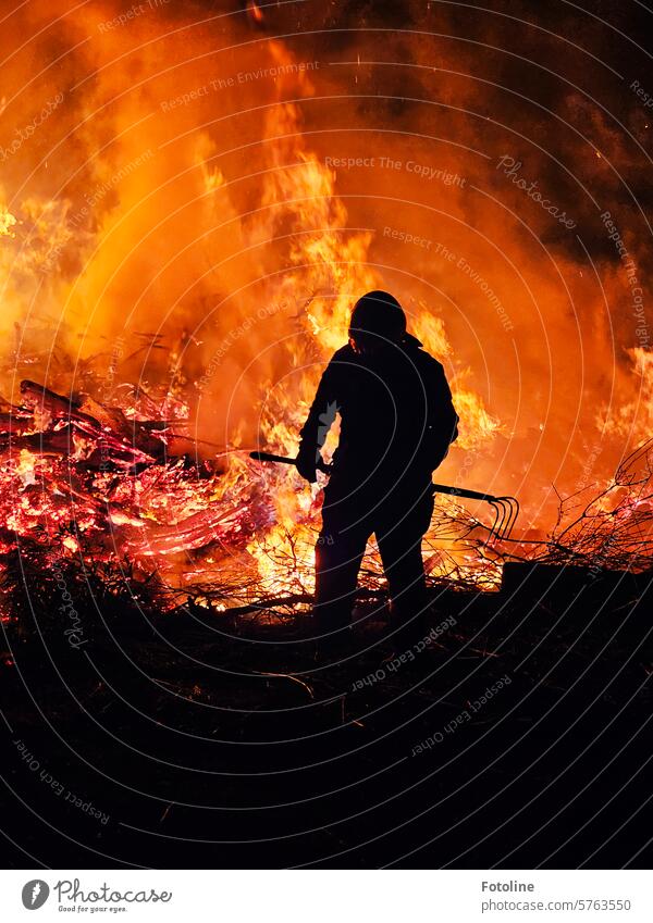 Ein Feuerwehrmann zieht mit einer Forke das Holz vom Rand in die Flammen, damit auch alle bösen Geister vertrieben werden. Hitze brennen heiß Wärme Feuerstelle