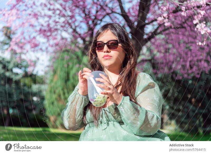 Junge Frau genießt ein Getränk in einem blühenden Park jung Sonnenbrille grünes Kleid Tasse schlürfen Blüte rosa pulsierend im Freien Frühling Natur Freizeit