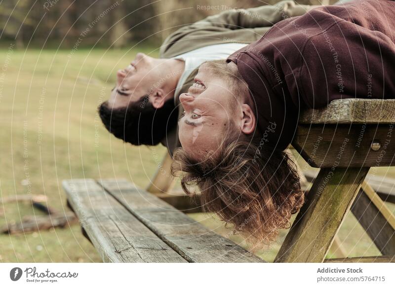 Umgekehrte Ansicht eines schwulen Paares, das zusammen lacht, während es sich auf einer Picknickbank zurücklehnt und einen spielerischen Moment im Freien genießt