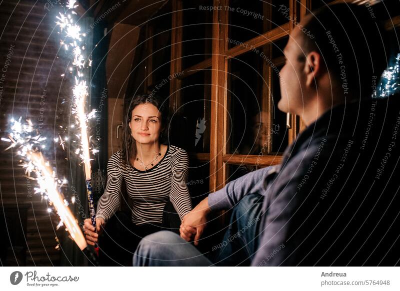 Ein Mann und ein Mädchen entzünden Wunderkerzen in einem Holzhaus Junge Lichter Fackel Spray Brandwunde Feuer schließen sitzen Abend Haus Baum Nacht Fenster