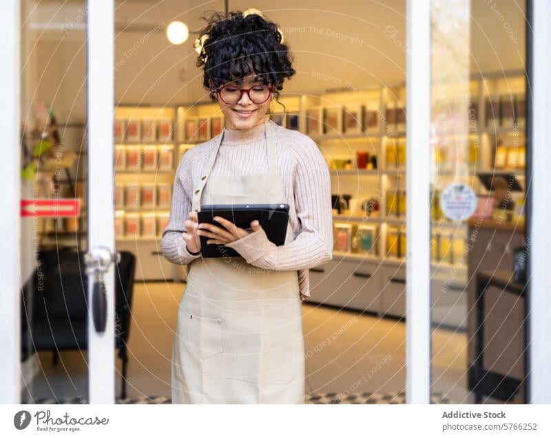 Lächelnde hispanische Geschäftsinhaberin mit Tablet im Schaufenster Frau Unternehmer Tablette digitales Gerät Ladenfront Boutique heiter Schürze Eingang