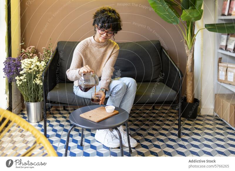 Lateinische Frau gießt kalten Kaffee im Haus mit stilvollem Dekor Gießen kaltes Brühen Glas geometrisch Muster Stock modern stylisch im Innenbereich trinken