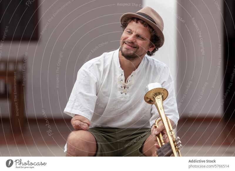 Entschlossener Musiker mit Trompete zu Hause einarmig Lächeln Mann Tatkraft praktizieren heimwärts Leidenschaft Sitzen lässig entspannt Hut Instrument Messing
