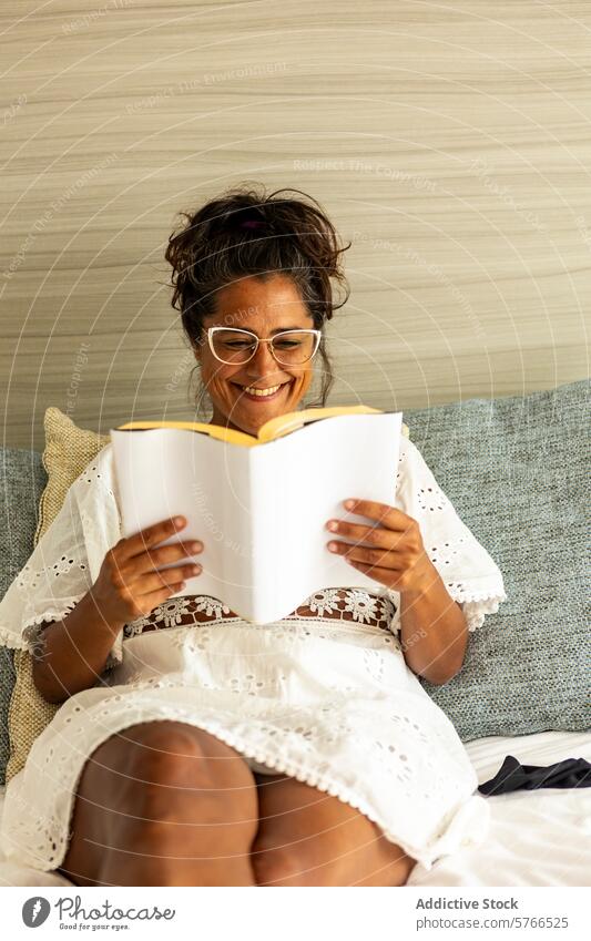 Fröhliche hispanische Frau mittleren Alters liest auf der Couch lesen Buch mittleres Alter Lächeln Liege gemütlich weiß Kleid Genuss Erholung Freizeit Hobby