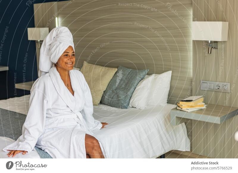 Entspannte hispanische Frau mittleren Alters im Bademantel auf einem Hotelbett sitzend Handtuch Bett Freizeit Komfort Gelassenheit Lächeln Inhalt entspannt