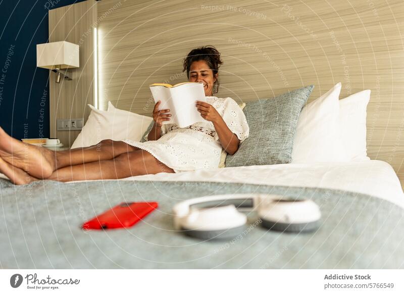 Hispanische Frau mittleren Alters lächelt, während sie in einem gemütlichen Schlafzimmer ein Buch liest lesen Bett Freizeit Zeit bequem Lügen Telefon Kopfhörer