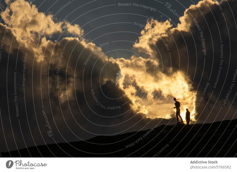 Die Silhouetten von zwei Personen stehen vor einem feurigen Sonnenuntergangshimmel auf einem Bergkamm, wobei die goldenen Wolken der Szene Dramatik verleihen