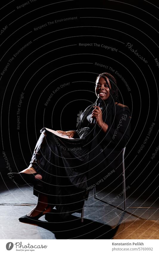 Vertikales Ganzkörperporträt einer lächelnden jungen Frau, die auf der Bühne in ein Mikrofon spricht, während sie in einem Stuhl mit Scheinwerferlicht sitzt
