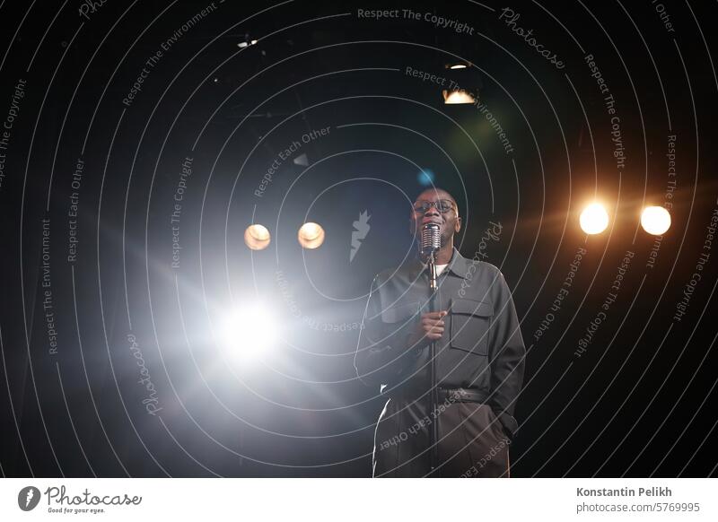 Weitwinkelaufnahme eines afroamerikanischen Mannes, der in ein Mikrofon spricht, auf einer Bühne mit Scheinwerferlicht steht und lächelnd den Raum kopiert