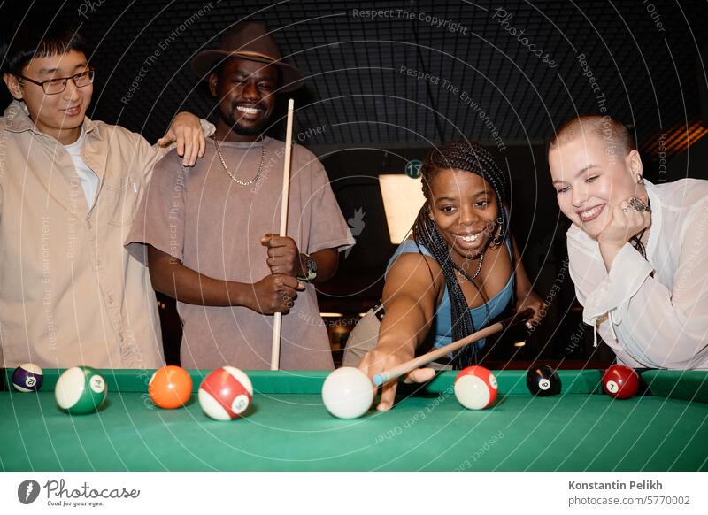 Multiethnische Gruppe lächelnder junger Leute, die zusammen mit einer schwarzen Frau, die eine Kugel mit einem Queue schlägt, Billard spielen Pool Mädchen Spiel