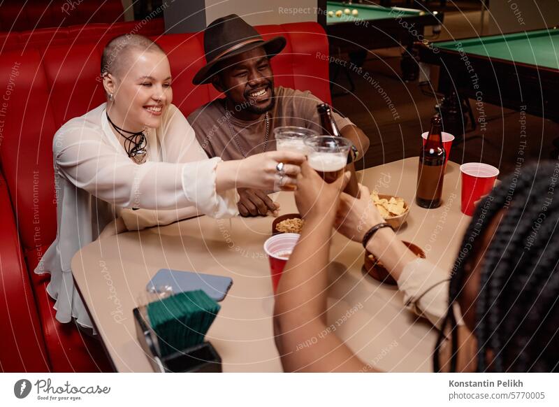 Eine multiethnische Gruppe von Freunden stößt mit Biergläsern an und sitzt jubelnd am Tisch Diner Café Menschengruppe trinken Klirren Glas Tasse Zuprosten Mann