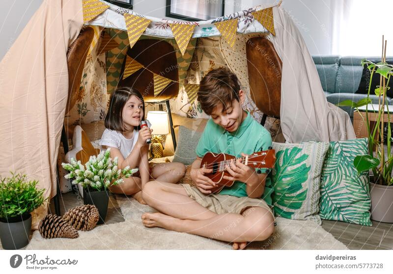 Glückliche Kinder spielen eine Ukulele und singen auf handgemachten Schutz Zelt im Wohnzimmer zu Hause Spielen Gitarre Gesang Musik Wartehäuschen Tipi Camping