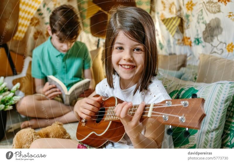 Porträt eines lächelnden kleinen Mädchens, das Ukulele spielt, während ein Junge auf einem handgefertigten Tipi zu Hause ein Buch liest Lächeln Frau Spielen