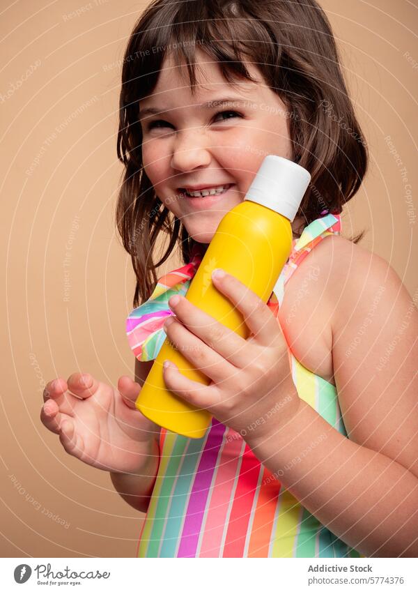 Junges Mädchen hält lächelnd eine gelbe Flasche Kind Lächeln Fröhlichkeit Kleid gestreift heiter farbenfroh beige Hintergrund Freude Beteiligung Griff hell Frau