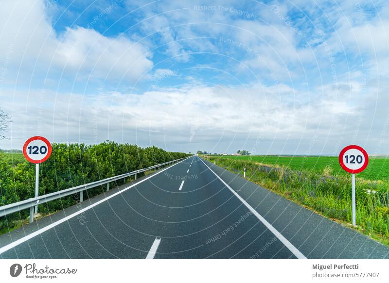 Blick auf eine gerade Autobahn, die im Hintergrund verschwindet, umrahmt von grünen Feldern unter einem blauen Himmel mit Wolken, mit zwei vertikalen Verkehrsschildern mit dem Limit von 120 km/h. Landschaft.