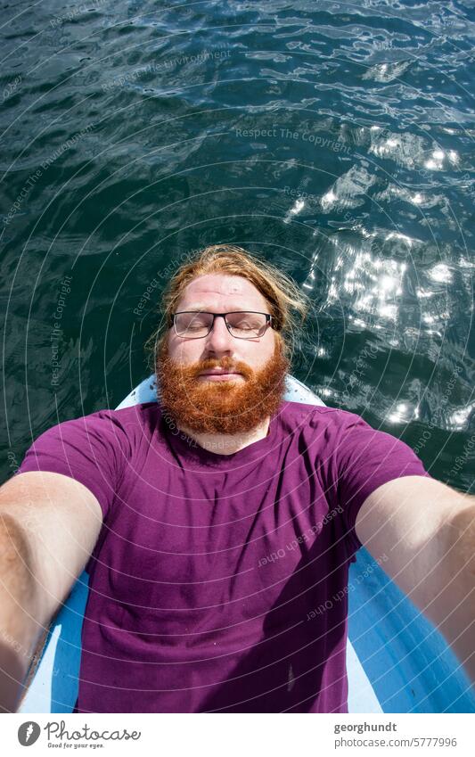 Mann mit rotem Bart und Brille lieg an der Spitze eines Ruderboots und genießt die Sonne. Dahinter ein See und die Spiegelung der Sonne. Der man hat ein violettes Shirt an, die Arme gehen nach vorn. Vielleicht fotografiert er sich selbst.