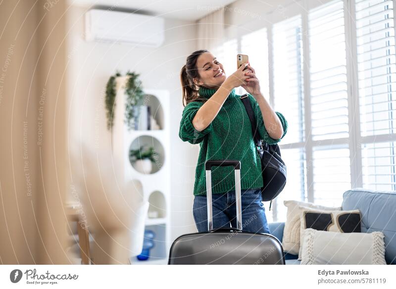 Junge Frau benutzt ein Mobiltelefon, während sie mit ihrem Koffer im Hotelzimmer steht Verpackung Bekleidung Ferien reisen Vorbereitung Menschen Gepäck