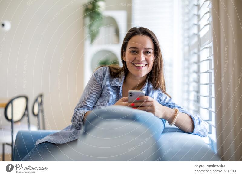 Lächelnde Frau mit Mobiltelefon auf dem Sofa im Wohnzimmer zu Hause Menschen eine Person Raum Erwachsener jung junger Erwachsener im Innenbereich Appartement
