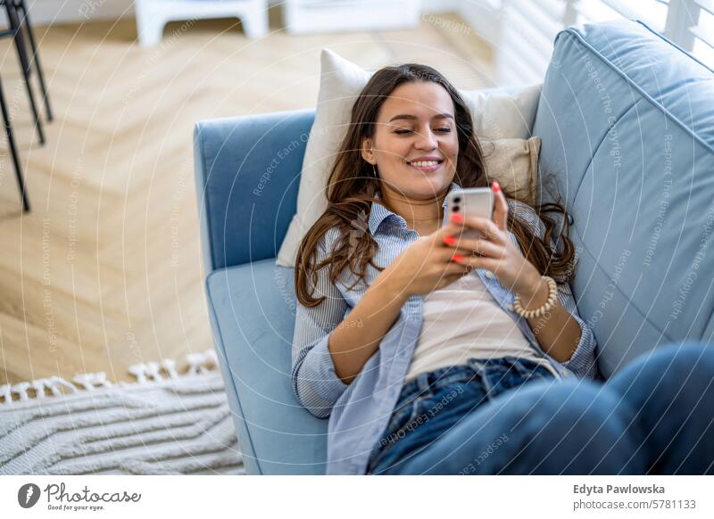 Lächelnde Frau mit Mobiltelefon auf dem Sofa im Wohnzimmer zu Hause Menschen eine Person Raum Erwachsener jung junger Erwachsener im Innenbereich Appartement