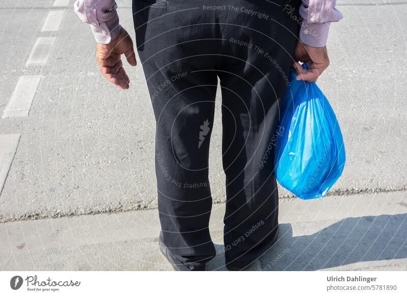 am Strassenrand stehende Person mit blauer Plastiktüte in der Hand.Abgebildet ist der Unterkörper mit schwarzer Hose und die anliegenden Hände Warten einkaufen