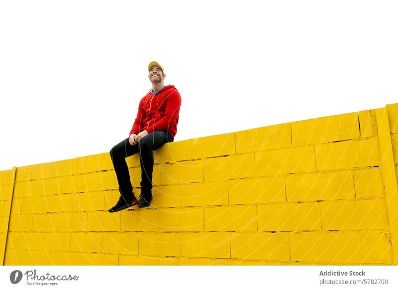 Mann mit rotem Kapuzenpulli sitzt auf einer hohen gelben Wand Sitzen lässig Kleidung Glück Freude pulsierend Farbe Kontrast hoch Baustein weiß Hintergrund