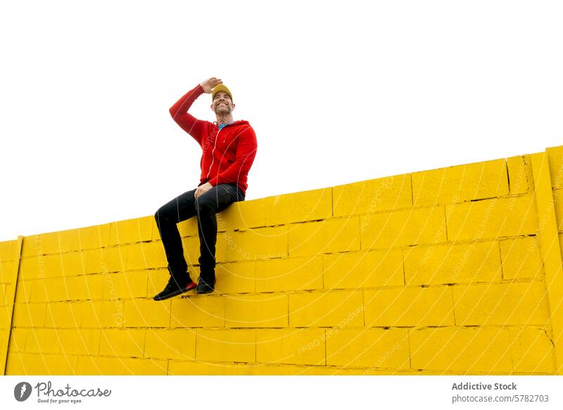 Mann, der auf einer hohen gelben Wand sitzt und die Augen abschirmt Sitzen Schutzschild Sonne heiter rot Kapuzenpulli pulsierend hell Farbe im Freien lässig