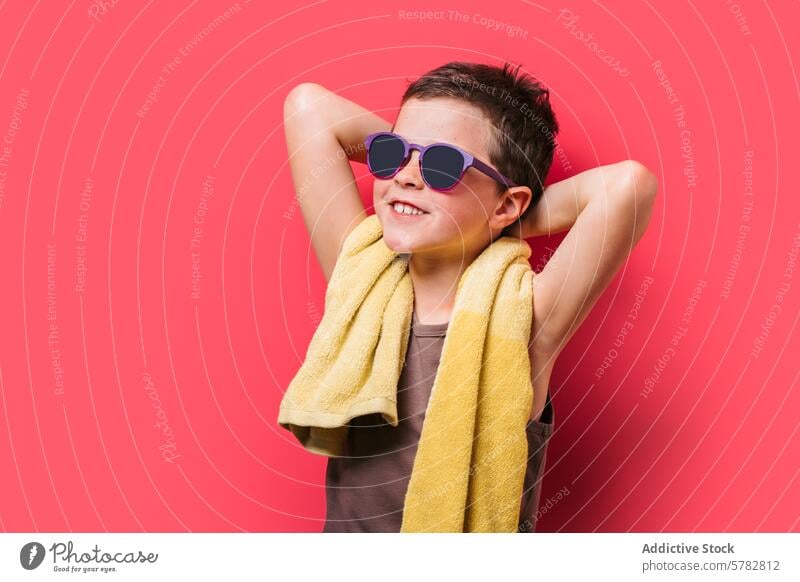 Glücklicher Junge mit Sonnenbrille genießt die Sommerstimmung Kind Lächeln heiter rosa Hintergrund gelbes Handtuch Freizeit Spaß lässig hell pulsierend Mode
