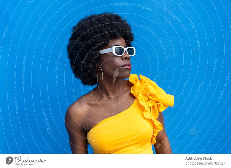 Modische Afro-Frau mit markantem blauem Hintergrund Afro-Look Mode Stil Sonnenbrille gelb Kleid pulsierend selbstbewusst Pose elegant schick trendy Sommer