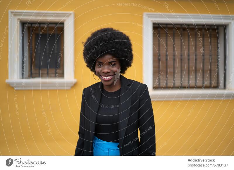 Lächelnde Afro-Frau in stilvollem urbanen Outfit Afro-Look Frisur selbstbewusst Pose gelb Wand Lifestyle pulsierend jung stylisch Kleidung Mode Porträt