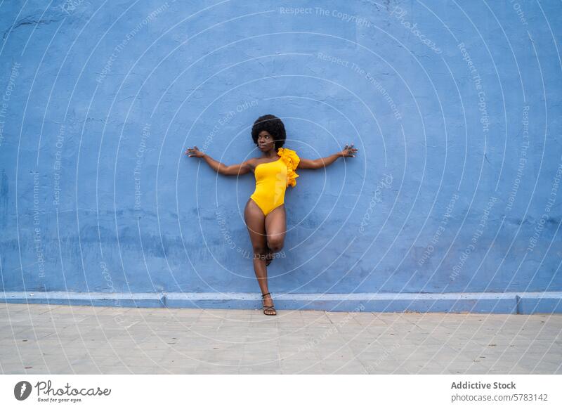 Lebhafte Afro-Frau vor blauer Wand in gelbem Outfit Afro-Look Lifestyle Mode Pose Selbstvertrauen Stil fett Textur Hintergrund Badeanzug Sommer urban trendy