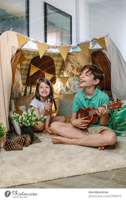 Glückliche Kinder spielen eine Ukulele und singen auf handgemachten Schutz Zelt im Wohnzimmer zu Hause Spielen Gitarre Lachen Gesang Musik Wartehäuschen Tipi