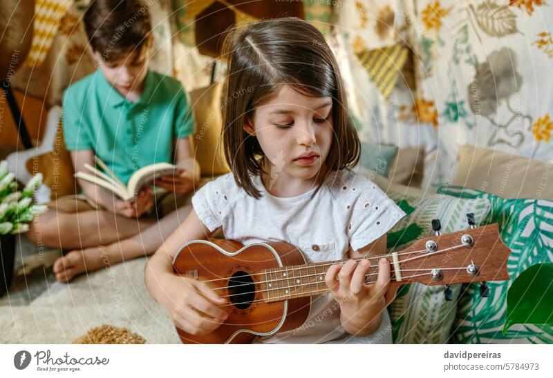 Porträt eines konzentrierten kleinen Mädchens, das Ukulele spielt, und eines Jungen, der auf einem handgefertigten Tipi zu Hause ein Buch liest Spielen Gitarre