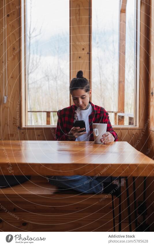 Lässige Frau mit Smartphone am Holztisch in einer Kabine Becher Fenster kariertes Hemd sitzend Beteiligung vertieft Gelassenheit lässig im Innenbereich