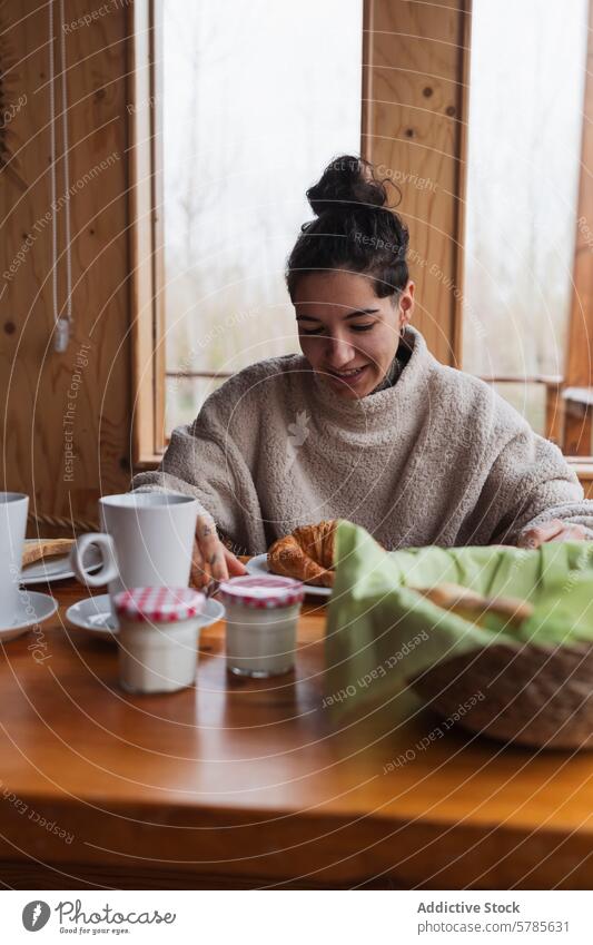 Frau genießt ein gemütliches Frühstück an einem kühlen Morgen Lächeln Holztisch Fenster genießen Szene Glück Croissant Kaffee Joghurt Brot Marmelade