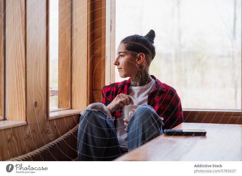 Nachdenkliche tätowierte Person mit Becher am Fenster Tattoo Kontemplation Gelassenheit Starrer Blick im Innenbereich jung nachdenklich kariertes Hemd Holz