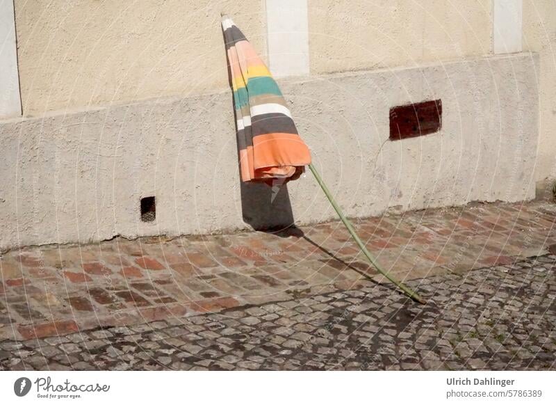 verbogener geschlossener mit blassfarbigen Streifen Sonnenschirm lehnt an einer Hauswand Kurios Humor lustig blasse Farben Strasse Schatten