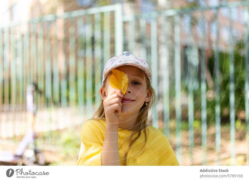 Mädchen mit einem gelben Blatt, das ein Auge bedeckt spielen Lächeln Verschlussdeckel Park hell verschwommener Hintergrund spielerisch Kindheit im Freien
