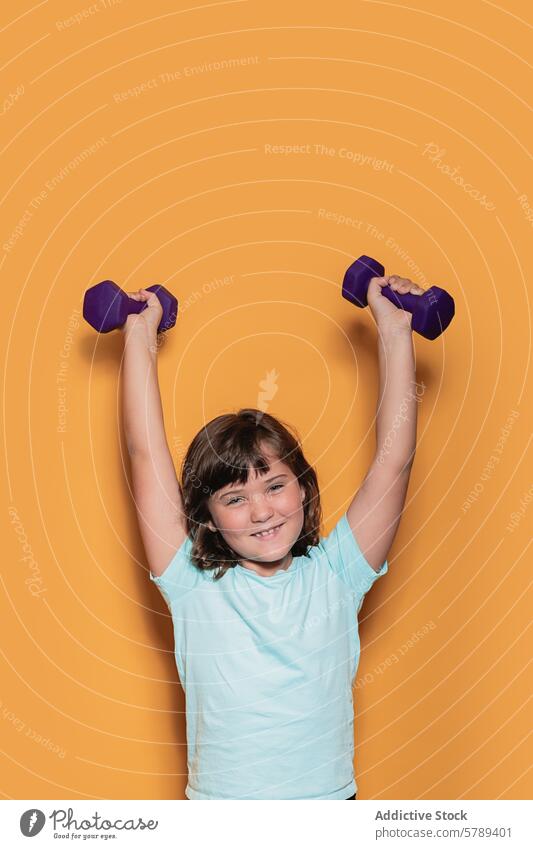 Fröhliches Kind hebt Gewichte auf orangefarbenem Hintergrund Fitness Gesundheit Sport Übung Mädchen Kurzhantel Heben Atelier Lächeln Freude Fröhlichkeit